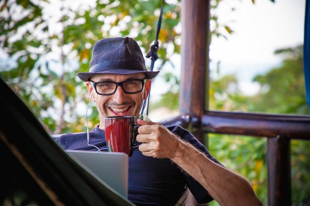 A man in a hammock uses a computer and drinks coffee