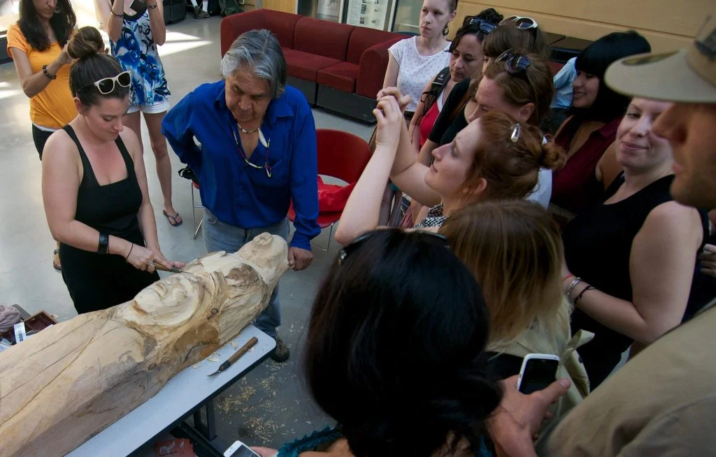 Isadore Charters (in blue) watches as a woman helps carve a totem pole at Simon Fraser University in Canada;